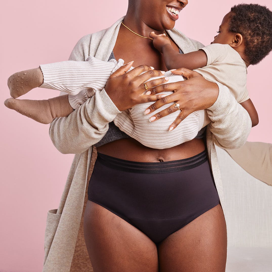 A mother is shown standing on a pink background smiling and holding her toddler while wearing a pair of Thinx for All Leaks bladder leak underwear in the style black Hi-Waist with a beige cardigan on